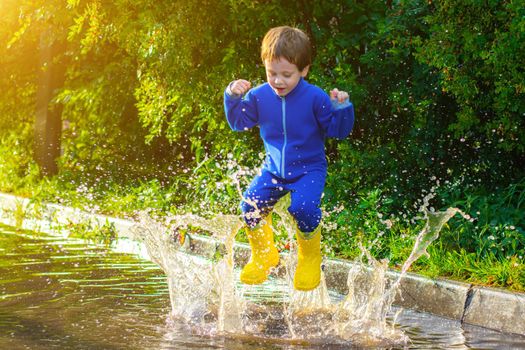 A happy boy in rubber boots jumps in puddles. The boy jumps in a puddle . Bad weather. puddles after rain. Childhood. A child in rubber boots. Summer walk. Happy child.