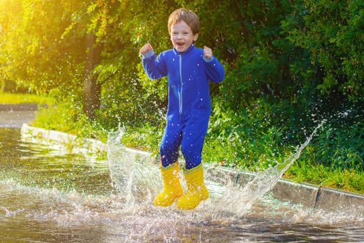 A happy boy in rubber boots jumps in puddles. The boy jumps in a puddle . Bad weather. puddles after rain. Childhood. A child in rubber boots. Summer walk. Happy child.
