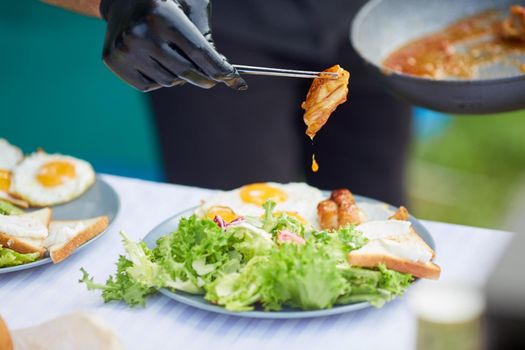 Selective focus of male hands in black gloves holding juicy meat with sauce drop using metal twizzers. Unrecognizable chef finishing dish, bread, salad and fried eggs on plates, table outdoors.