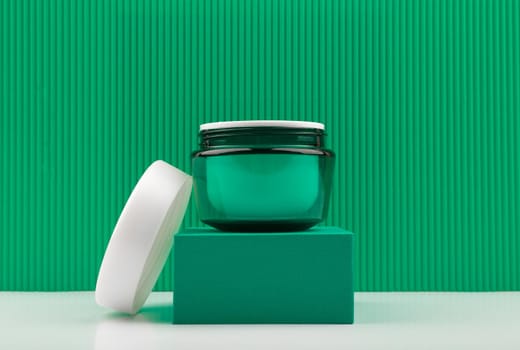 Close up of face cream, mask or scrub in green opened jar on green podium against green background with stripes. Concept of luxury organic cosmetics