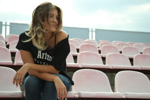 urban portrait of a flirty beautiful woman in black t-shirt and jeans at the stadium, sitting on a bench.