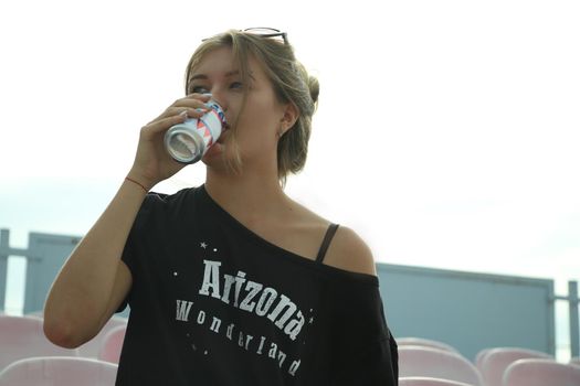 urban portrait of a flirty beautiful woman in black t-shirt and jeans at the stadium, sitting on a bench, drinking from a metal jar.