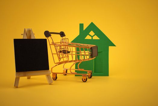 Property concept with house, small chalkboard and shopping cart over yellow background and free copy space for text