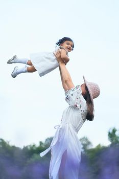 Young loving mother wearing straw hat enjoying time with little baby daughter, playing with laughing kid. Stunning lady wearing dress throwing child up, blooming lavender field on background.