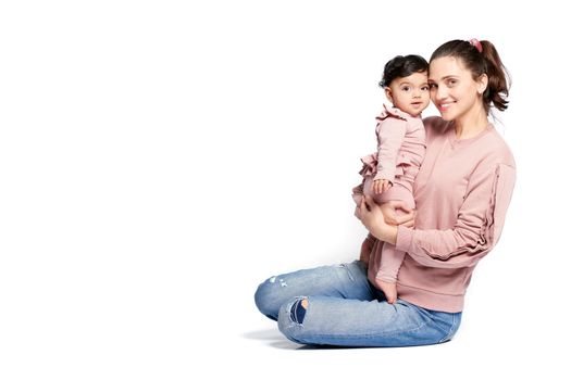 Side view of mother with smiling baby daughter looking at camera isolated on white studio background. Portrait of young woman holding sweet adorable child in arms while sitting on floor.