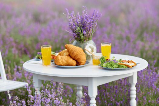 White wooden table served with delicious croissants, glasses of orange juice, honey jar and fresh appetizers for two and vase with lavender bouquet. Amazing decoration in blooming lavender field.