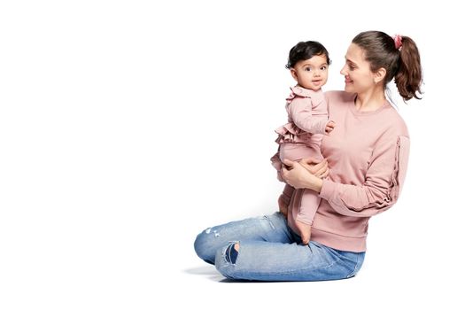 Portrait of mother with smiling baby daughter isolated on white studio background. Side view of young woman holding sweet adorable child in arms while sitting on floor, happy kid looking at camera.