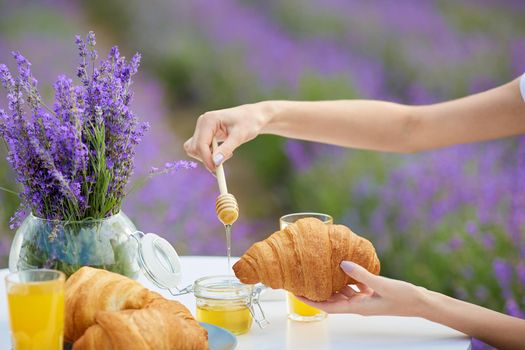 Side view of female hands putting honey using wooden spoon on fresh croissants, vase with lavender bouquet and glasses of orange juice. Beautiful decorated table in blooming lavender field.