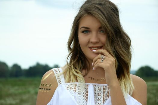 portrait of a happy flirty beautiful blonde woman in white blouse in the field. Temporary tattoo. Drawings on body. hippie. Nature loving.