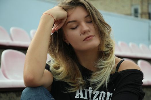 urban portrait of a flirty beautiful woman in black t-shirt and jeans at the stadium, sitting on a bench.