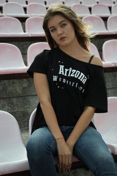 urban portrait of a flirty beautiful woman in black t-shirt and jeans at the stadium, sitting on a bench.