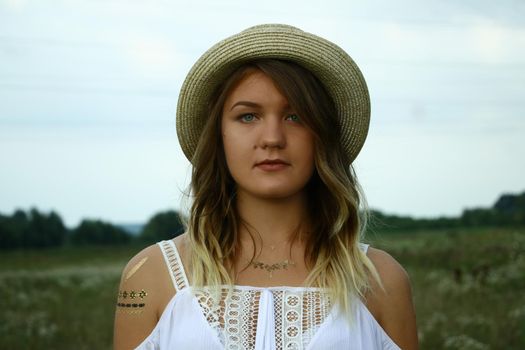 portrait of a pensive beautiful blonde woman in white blouse and straw hat in the field. Temporary tattoo. Drawings on body. hippie. Nature loving.