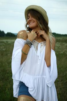 portrait of a happy flirty beautiful blonde woman in white blouse and straw hat in the field. Temporary tattoo. Drawings on body. hippie. Nature loving.