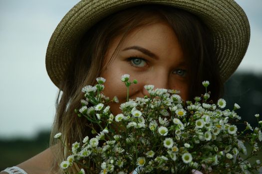 portrait of a happy flirty beautiful blonde woman in white blouse and straw hat in the field. Carrying a bouquet of daisies. Temporary tattoo. Drawings on body. hippie. Nature loving.