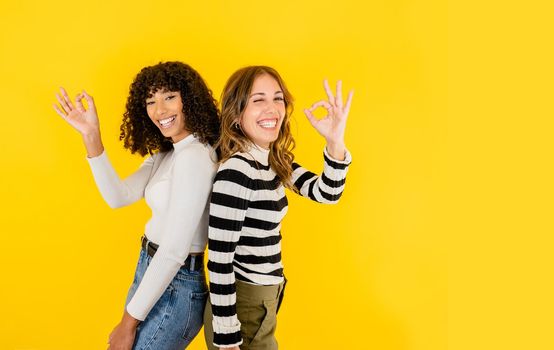 Two multiracial young beautiful models posing with ok gesture in toothy smile isolated on yellow background for copy space. Concept of positive mood, work success, and happiness enjoying your life