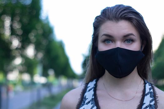 Pretty young blonde woman in medical black face mask. Wearing a t-shirt and jeans shorts. in a park. modern reality. covid-19 concept. copy space. High quality photo