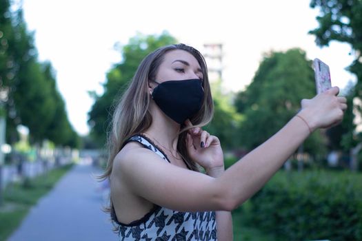 Pretty young blonde woman in medical black face mask. Wearing a t-shirt and jeans shorts. in a park. covid-19 concept. copy space.