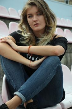 urban portrait of a flirty beautiful woman in black t-shirt and jeans at the stadium, sitting on a bench.