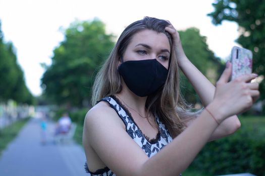 Pretty young blonde woman in medical black face mask. Wearing a t-shirt and jeans shorts. in a park. covid-19 concept. copy space.