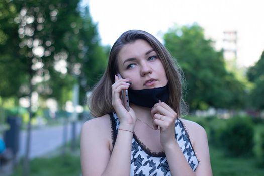 Pretty young blonde woman in medical black face mask. Wearing a t-shirt and jeans shorts. in a park. covid-19 concept. copy space.