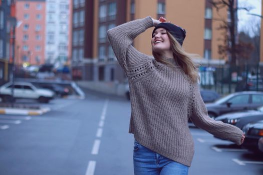 urban portrait of a happy flirty beautiful woman in knitted brown sweater, jeans and black hat. in the parking lot.