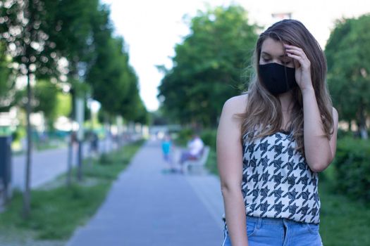 Pretty young blonde woman in medical black face mask. Wearing a t-shirt and jeans shorts. in a park. modern reality. covid-19 concept. copy space. High quality photo