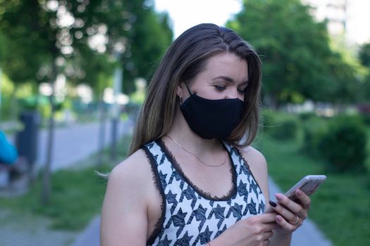 Pretty young blonde woman in medical black face mask. Wearing a t-shirt and jeans shorts. in a park. covid-19 concept. copy space.