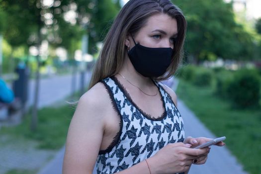 Pretty young blonde woman in medical black face mask. Wearing a t-shirt and jeans shorts. in a park. covid-19 concept. copy space.