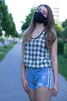 Pretty young blonde woman in medical black face mask. Wearing a t-shirt and jeans shorts. in a park. modern reality. covid-19 concept. copy space. High quality photo