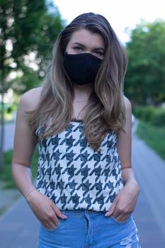 Pretty young blonde woman in medical black face mask. Wearing a t-shirt and jeans shorts. in a park. modern reality. covid-19 concept. copy space. High quality photo