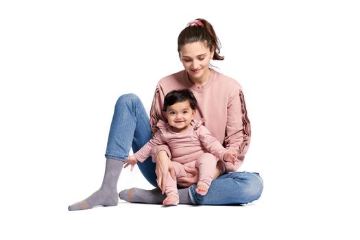 Portrait of cute mother with smiling baby daughter isolated on white studio background. Young attractive woman holding sweet adorable child on leg while sitting on floor, happy childhood concept.