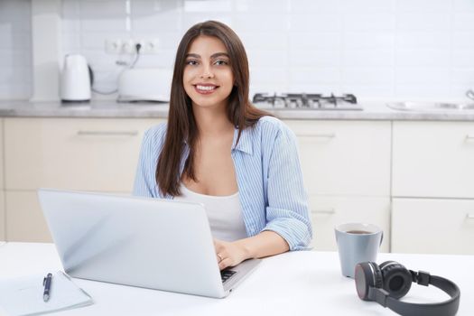 Close up of young smiling beautiful woman using laptop for working or studying at home. Concept of process working or studying in kitchen.