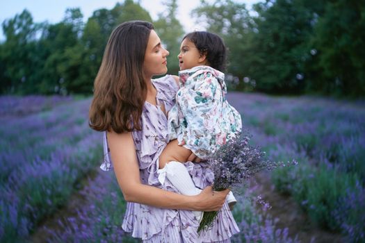 Side view of young loving woman posing with baby girl on hands in summer lavender field. Happy smiling mother wearing dress carrying beautiful bouquet of purple flowers. Concept of nature beauty.