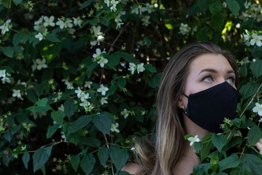 Pretty young blonde woman in medical black face mask. in a bush with little white flowers. modern reality. covid-19 concept. copy space. High quality photo