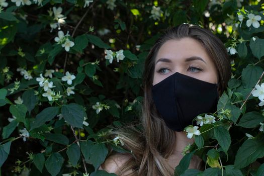 Pretty young blonde woman in medical black face mask. in a bush with little white flowers. modern reality. covid-19 concept. copy space. High quality photo