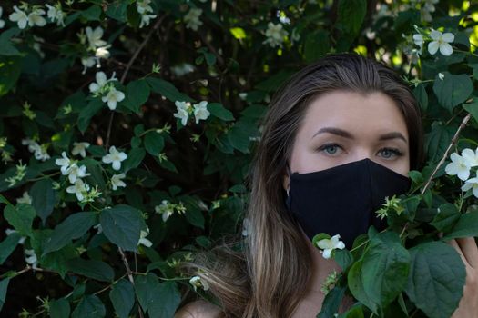 Pretty young blonde woman in medical black face mask. in a bush with little white flowers. modern reality. covid-19 concept. copy space. High quality photo