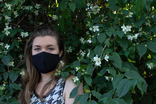 Pretty young blonde woman in medical black face mask. in a bush with little white flowers. modern reality. covid-19 concept. copy space. High quality photo