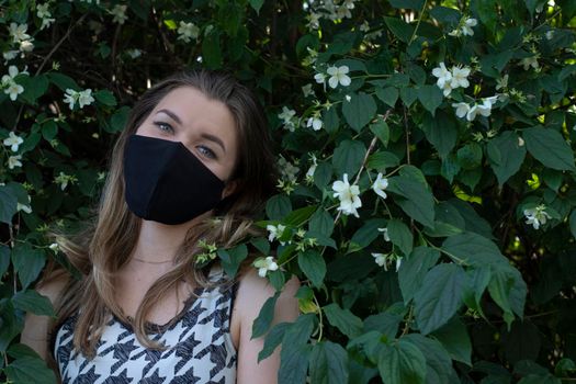 Pretty young blonde woman in medical black face mask. in a bush with little white flowers. modern reality. covid-19 concept. copy space. High quality photo