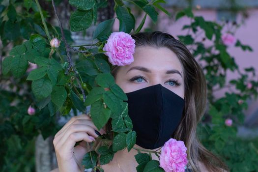 Pretty young blonde woman in medical black face mask. near a bush with pink flowers. modern reality. covid-19 concept. copy space. High quality photo