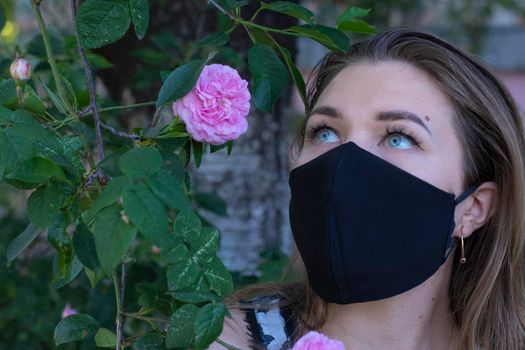 Pretty young blonde woman in medical black face mask. near a bush with pink flowers. modern reality. covid-19 concept. copy space. High quality photo