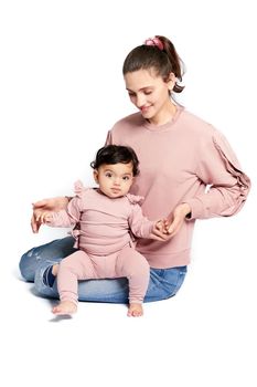 Portrait of cute mother with daughter looking at camera isolated on white studio background. Young attractive woman sitting on floor and helping sweet adorable child to stand, happy childhood concept.