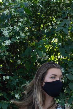 Pretty young blonde woman in medical black face mask. in a bush with little white flowers. modern reality. covid-19 concept. copy space. High quality photo