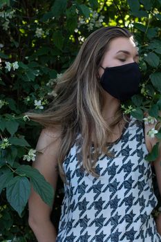 Pretty young blonde woman in medical black face mask. in a bush with little white flowers. modern reality. covid-19 concept. copy space. High quality photo
