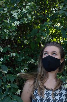 Pretty young blonde woman in medical black face mask. in a bush with little white flowers. modern reality. covid-19 concept. copy space. High quality photo