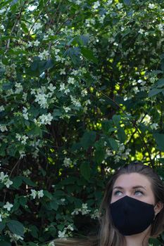 Pretty young blonde woman in medical black face mask. in a bush with little white flowers. modern reality. covid-19 concept. copy space. High quality photo