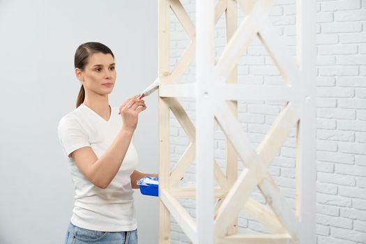 Young brunette woman holding brush and painting wooden rack in empty room. Concept of repair at home.