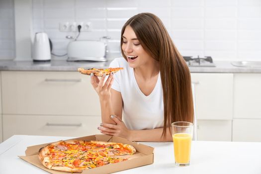 Pretty woman eating tasty pizza and drinking juice in kitchen. Concept of pizza time at home.