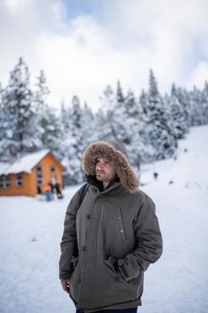 Hooded man standing with a beautiful snowy forest and small cabin as background. Portrait of happy man surrounded by snowy pine trees. Adventurous winter holiday