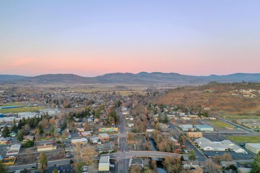 Peaceful town surrounded by trees with mountains as background during beautiful sunset. Aerial view of roads surrounded by autumn-colored nature. Rural town landscape