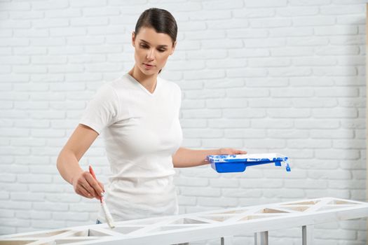 Young beautiful brunette holding white paint and painting wooden rack. Concept of improvement rack at home.